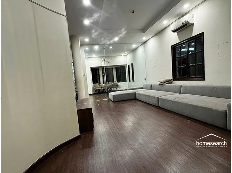 This living area features dark wood-look flooring and neutral walls, creating a clean, modern aesthetic. It is equipped with a wall-mounted AC unit and a large grey sectional sofa, with a glass partition separating it from an adjacent room to maintain an open, airy flow.