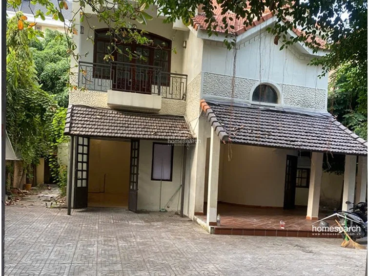 The home features a traditional facade with a mix of smooth stucco and textured stone detailing, complemented by dark wood-framed windows and doors. The ground level offers a spacious, covered outdoor terrace that flows directly onto a paved courtyard, while the upper floor includes a private balcony with ornate metal railings.