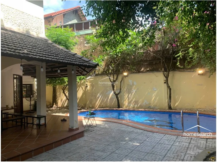 This perspective highlights the covered patio area, which is paved with terracotta tiles and supported by sturdy white columns, creating a perfect space for outdoor dining or relaxation. The area is illuminated by hanging pendant lights and overlooks the swimming pool, offering a tranquil view of the lush, tree-lined garden.