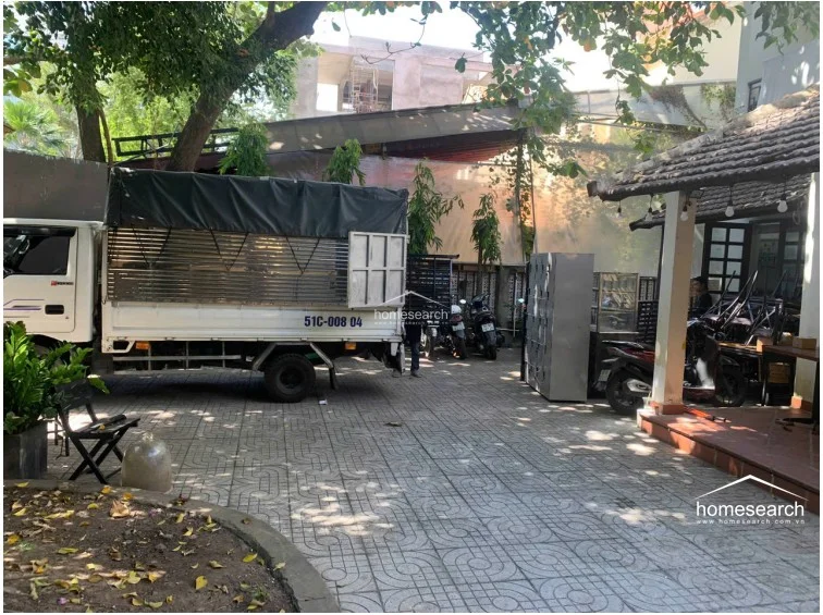 This view captures a side yard or driveway area currently used for storage and vehicle parking, featuring a large transport truck and various loose items. The space is paved with concrete tiles and is bordered by a mix of mature trees and a traditional tiled-roof structure, suggesting a property with significant outdoor utility space.