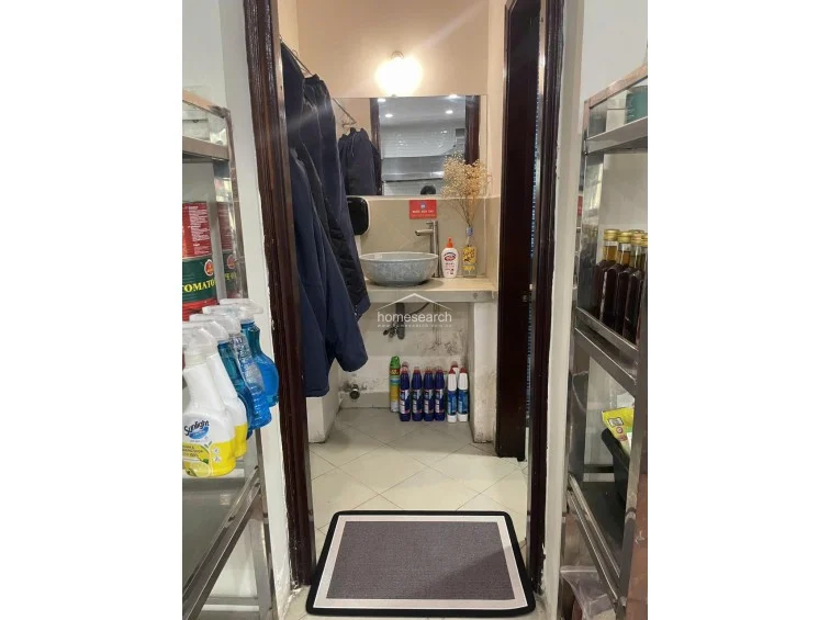 This small, functional washroom or utility transition area is framed by dark wood trim and features a vessel sink on a stone vanity. The space is used for storage of cleaning supplies and personal items, with a simple overhead light fixture and neutral floor tiles.