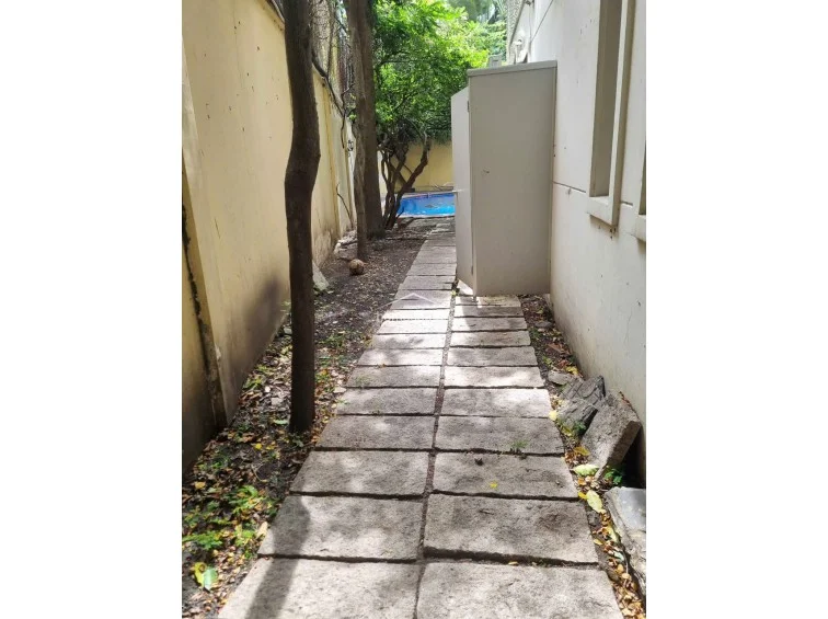 This narrow garden path is lined with large stone pavers and leads toward a glimpse of a swimming pool in the distance. The area is flanked by a high perimeter wall on one side and the exterior of the building on the other, with an upright storage cabinet positioned along the walkway.