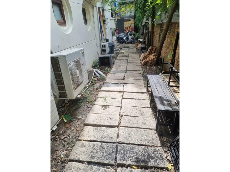 This exterior walkway features a stone-paved path running alongside the building, which is equipped with multiple external AC compressor units. The area is functional but currently cluttered with construction materials and outdoor furniture, highlighting a need for improved maintenance and organization in this side-yard space.