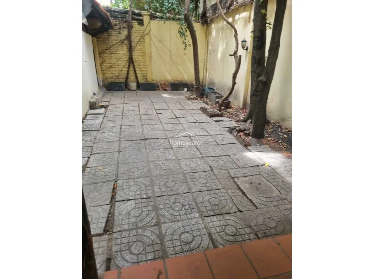 This outdoor area features a paved patio with patterned concrete tiles, enclosed by high yellow brick walls that provide privacy. The space is shaded by mature trees and includes several empty planters, suggesting a low-maintenance garden area that could be revitalized with landscaping.