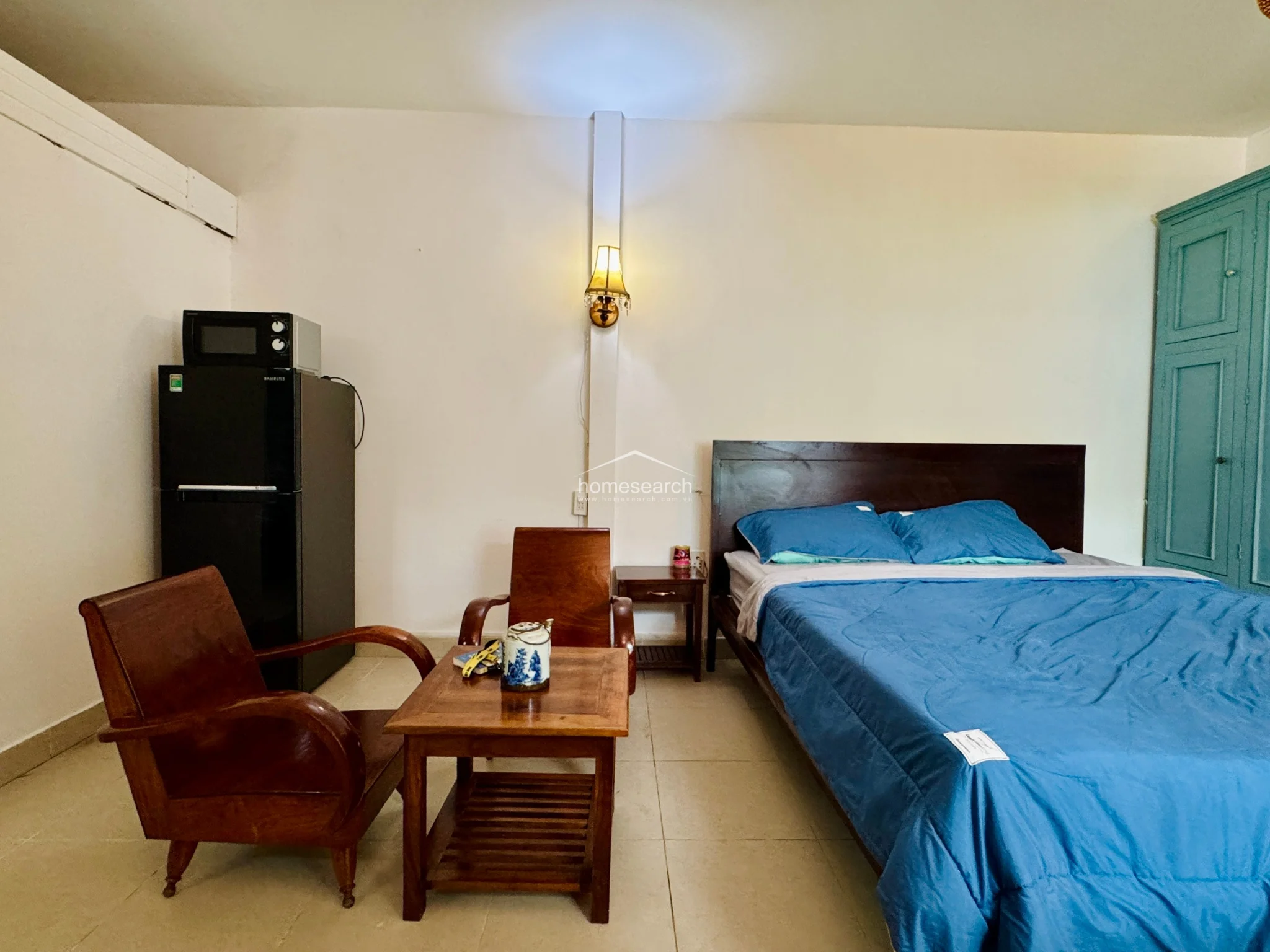 The sleeping area is anchored by a dark wood bed frame dressed in bright blue bedding, positioned adjacent to a large, matching green built-in wardrobe. A small wooden side table and a pair of traditional wooden armchairs with a low tea table create a cozy seating nook. The room maintains a consistent aesthetic with warm, wall-mounted sconce lighting and minimalist decor.