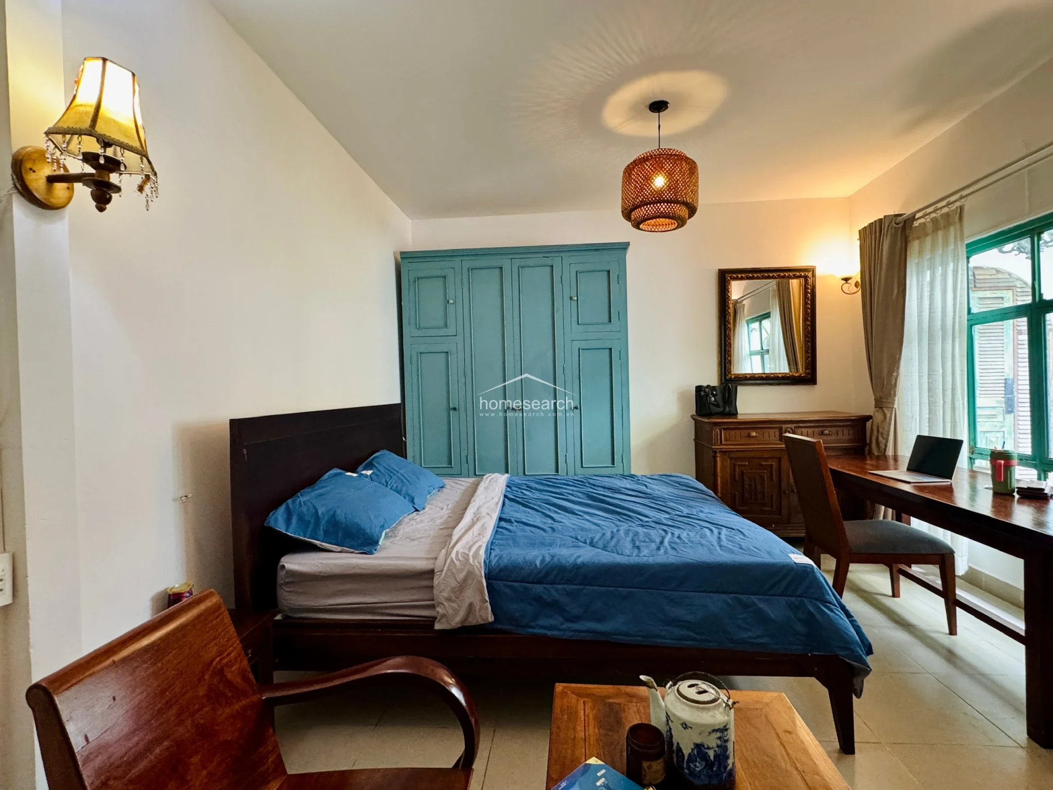 This view highlights the functional flow of the studio, showing the relationship between the sleeping quarters and the dedicated workspace by the window. The room is well-maintained, featuring a classic wooden desk and chair set that takes advantage of the natural light. The overall atmosphere is calm and functional, with a neutral color palette punctuated by the bold green architectural accents.