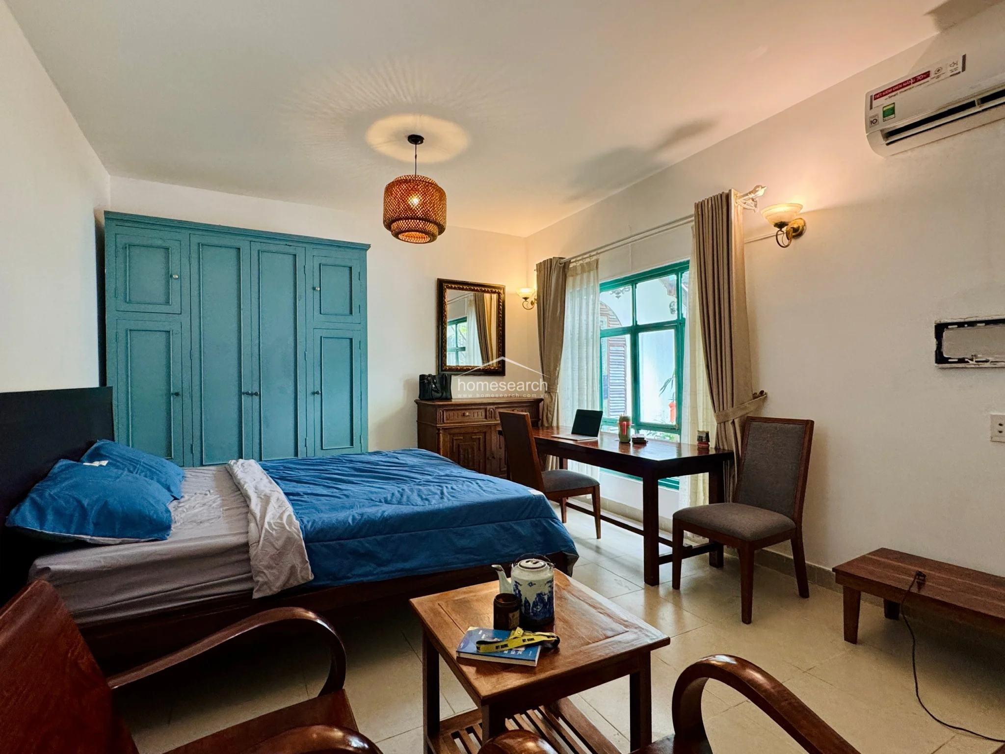 This angle emphasizes the storage and utility aspects of the studio, including the large green wardrobe and a separate wooden dresser topped with a decorative mirror. The layout is efficient, placing the sleeping area near the kitchen and laundry zone, which is tucked away behind a partition. The combination of wood furniture and white walls creates a balanced, clean look throughout the living space.