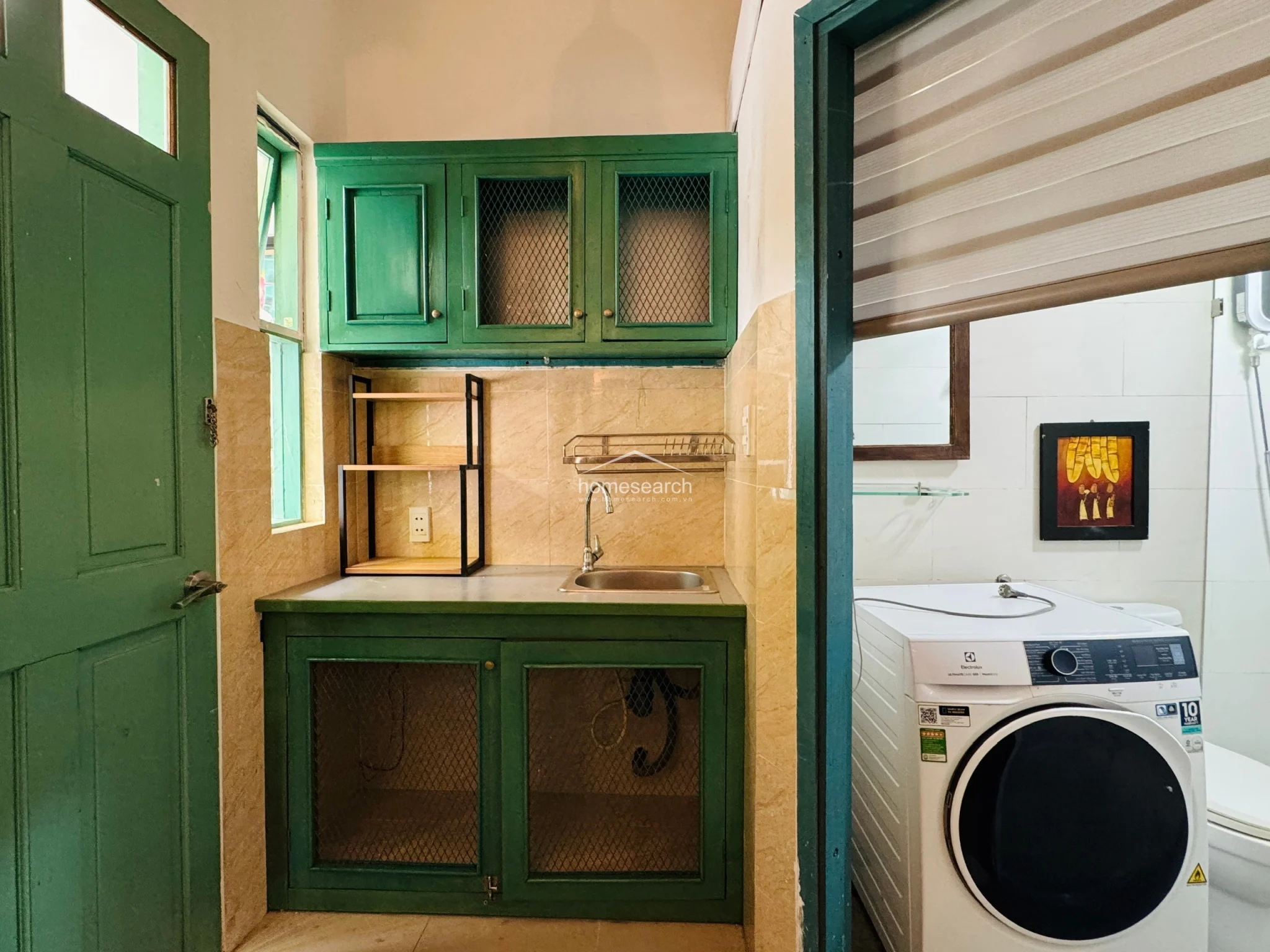 The kitchen area is designed with a practical, compact layout featuring vibrant green cabinets with wire-mesh detailing and a neutral tiled backsplash. Adjacent to the sink area, a doorway leads directly into a bright, tiled bathroom equipped with a modern front-loading washing machine and a shower area. The space is well-maintained, utilizing a cohesive color scheme that bridges the kitchen and utility zones.