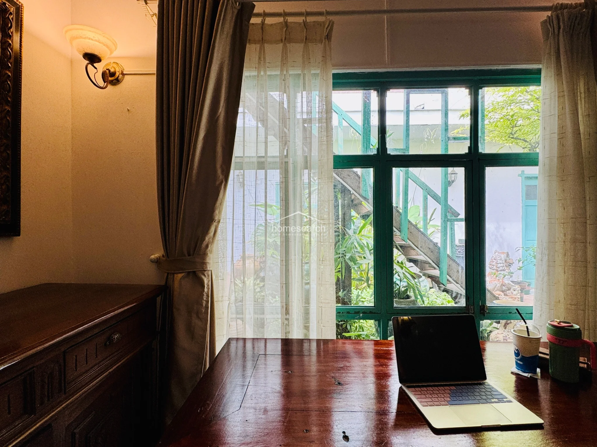 This close-up view highlights a cozy workspace featuring a rich, dark-stained wooden desk positioned against a large window with green frames. The space is accented by a warm wall-mounted sconce and soft, neutral-toned curtains, creating an inviting and tranquil atmosphere with a view of an outdoor staircase and lush greenery.