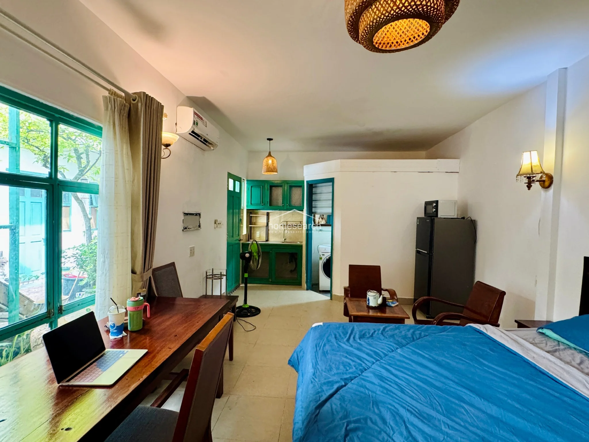 This studio apartment features an open-plan layout that seamlessly integrates a sleeping area, a workspace, and a compact kitchenette. The space is finished with light-colored tile flooring and white walls, accented by vibrant green painted door and cabinet frames that add a distinct character. Large windows with green frames provide ample natural light and a view of lush greenery, while a wall-mounted AC unit and woven pendant lighting ensure comfort and style.