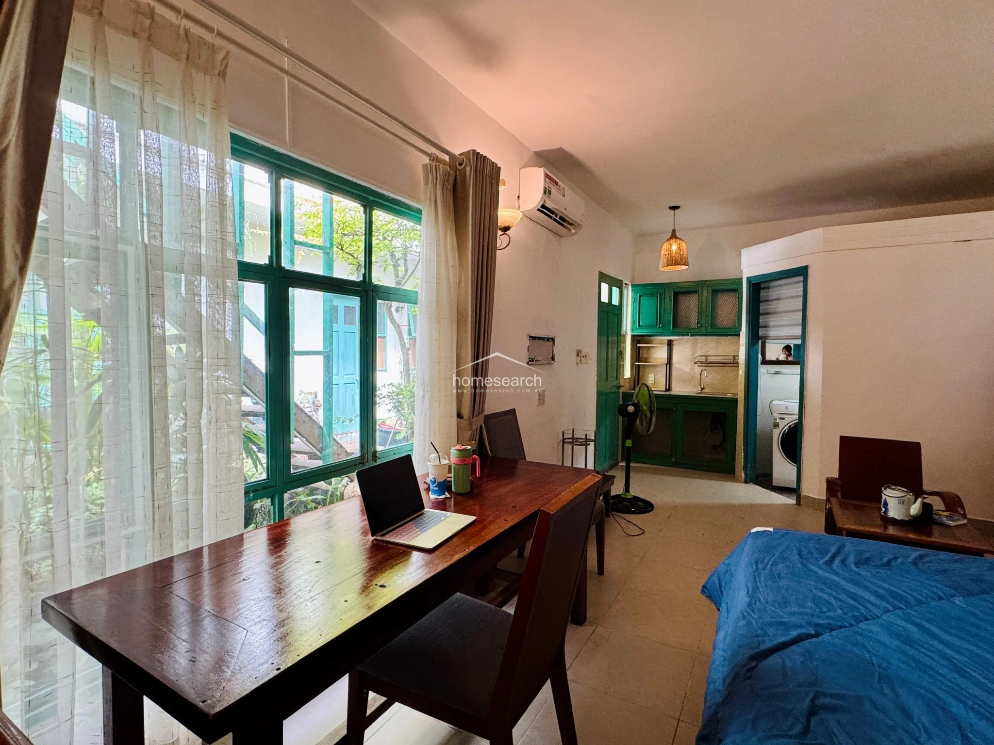 This studio apartment layout efficiently integrates a bedroom, dining area, and kitchen into an open-concept flow. The room features light-colored tile flooring, white walls, and a prominent green-framed window that provides ample natural light and a view of an outdoor garden area. The kitchen is compact with green cabinetry, while a wall-mounted AC unit and a ceiling-mounted pendant light ensure comfort and functional illumination.