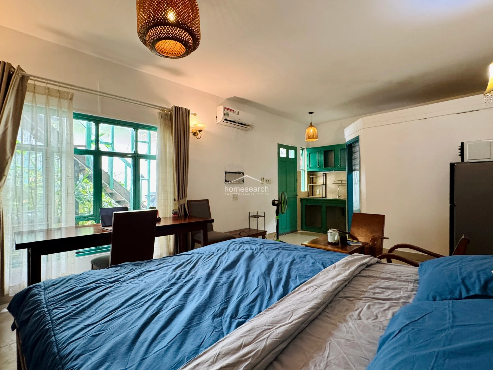 This perspective showcases the kitchenette area, which is equipped with green cabinetry and a small laundry nook visible through a doorway. The studio is well-equipped with modern essentials, including a refrigerator and microwave, while the large window ensures the entire space feels bright and airy. The property appears to be in good condition, offering a comfortable and well-organized living environment.
