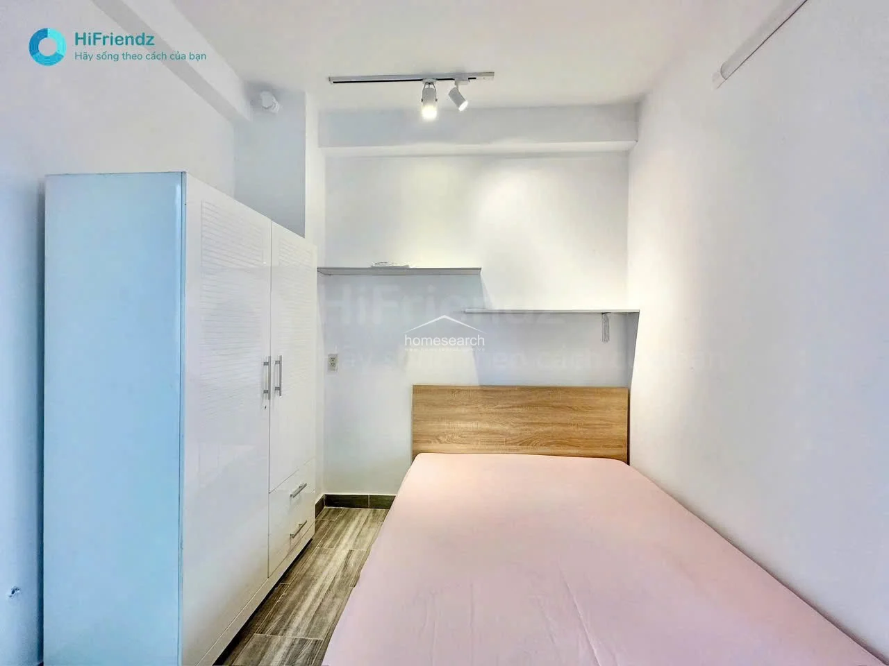 This bedroom features a minimalist design with white walls and wood-patterned tile flooring, creating a clean and bright atmosphere. It includes a large white wardrobe for storage, a bed with a light pink cover, and a wooden headboard, all illuminated by modern track lighting.