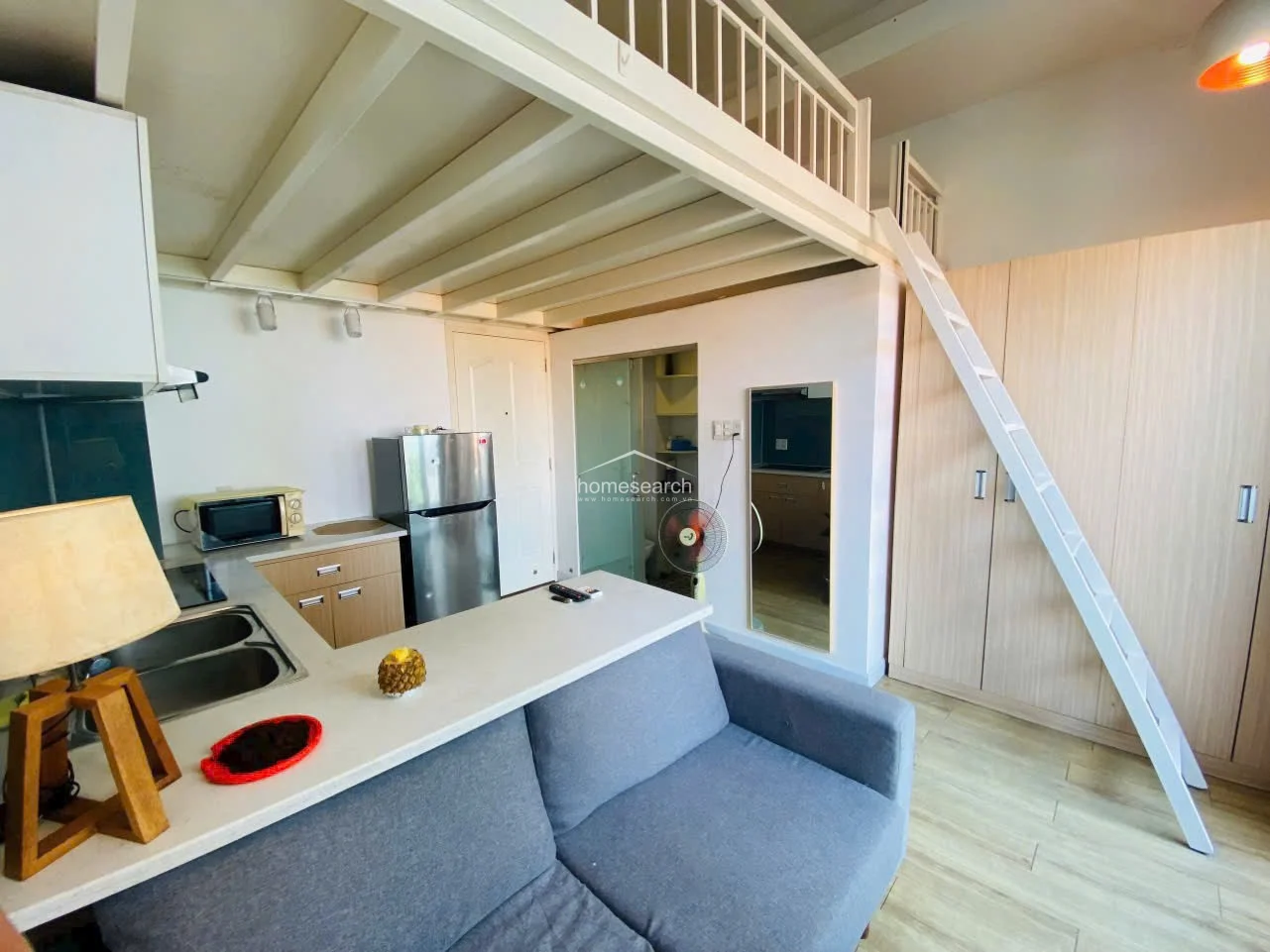 From this angle, the living space showcases the clever use of a loft mezzanine, which creates a distinct separation between the sleeping and living zones. The kitchen area is neatly integrated with cabinetry and a breakfast bar, while a full-length mirror adds depth to the room.