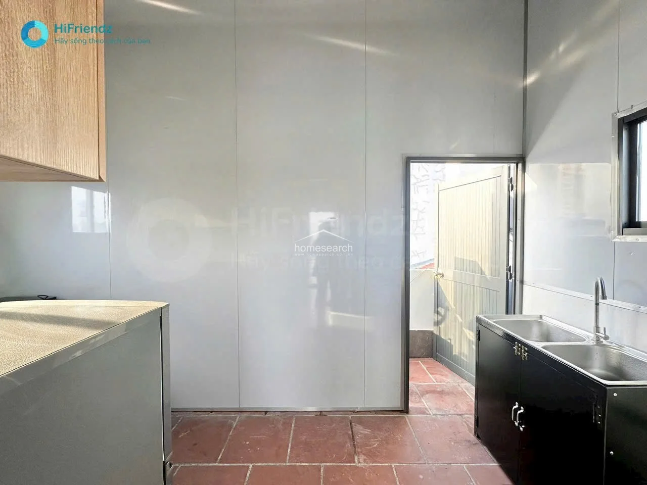 This utility or kitchen area features red-toned tile flooring and white wall paneling, offering a durable and easy-to-clean workspace. It includes a stainless steel double sink and a door leading directly to an outdoor space, providing good ventilation and natural light.