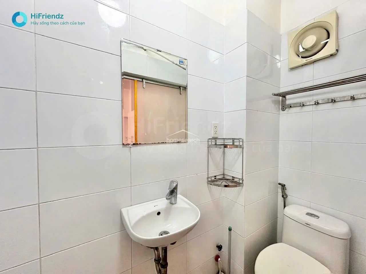 This compact bathroom features white wall tiles throughout and a standard white pedestal sink paired with a simple wall-mounted mirror. Ventilation is provided by an integrated exhaust fan, and the space includes a basic toilet and a stainless steel corner shelf for storage.