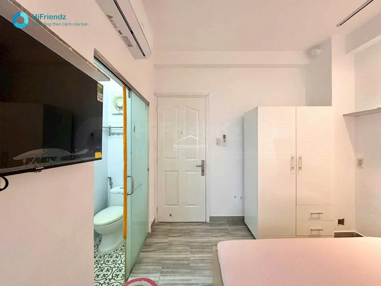 The bedroom area is equipped with a wall-mounted television, a split-type AC unit, and a large white wardrobe for storage. It connects directly to a small, tiled balcony area, providing natural light and a transition to the outdoors.