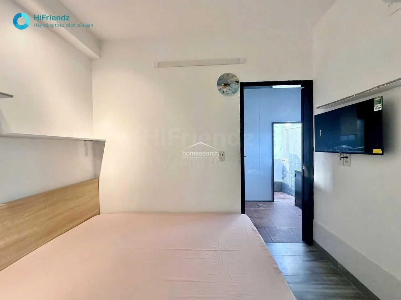 This bedroom space is minimalist and bright, featuring a bed with a light wood headboard and a wall-mounted TV. The room maintains a consistent aesthetic with the rest of the unit, utilizing light wood flooring and white walls to create an airy, comfortable atmosphere.