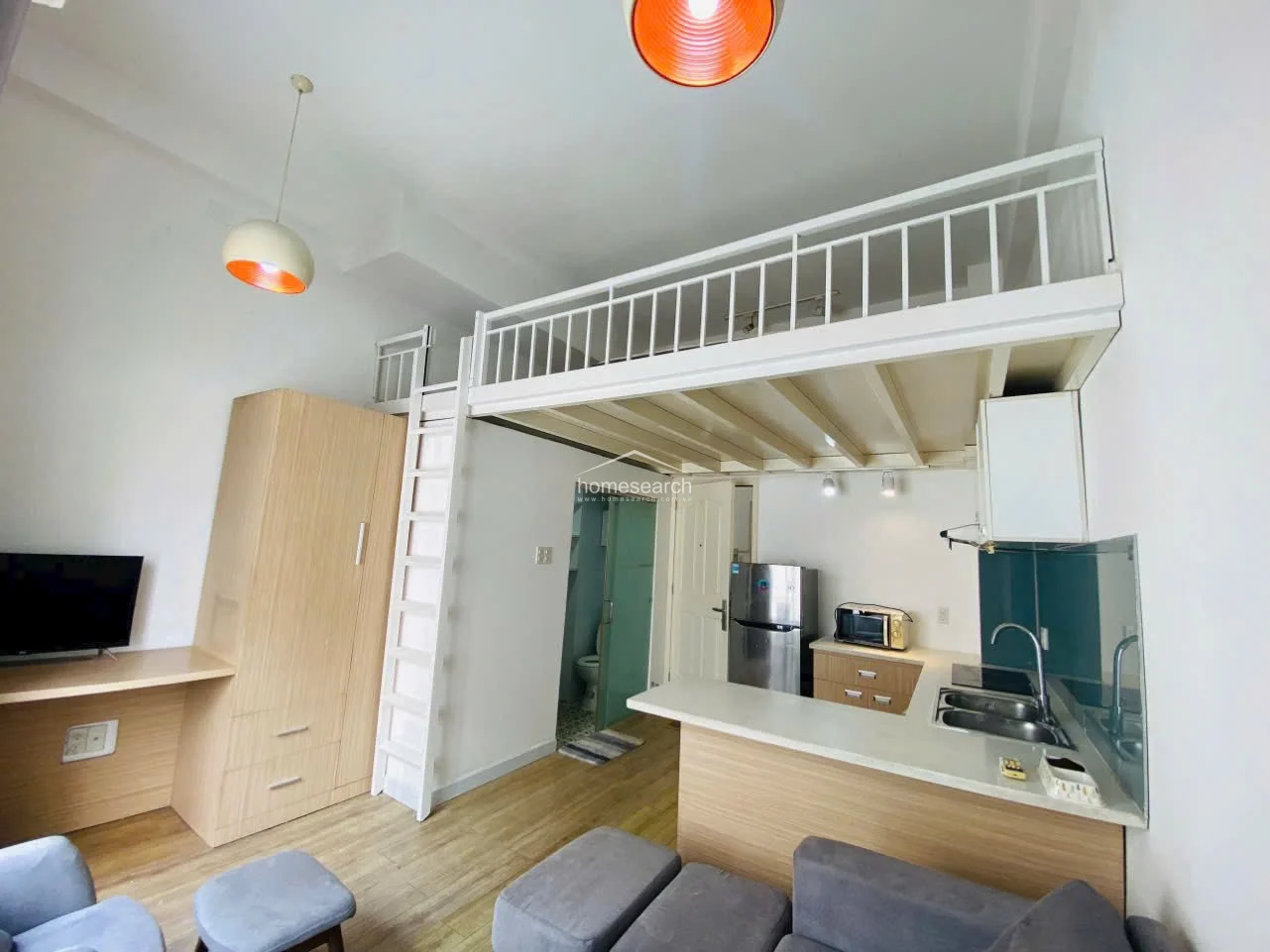 This studio apartment utilizes a clever loft layout to maximize space, featuring a sleeping area above and a living/kitchen area below. The space is furnished with a grey sofa, a light wood wardrobe, and a functional kitchen counter, while large windows provide ample natural light and a view of the outdoors.