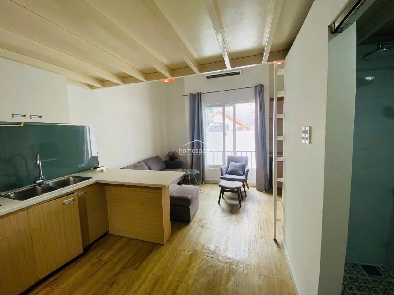 This view highlights the open-plan flow of the studio, showing the connection between the kitchen, living area, and the loft ladder. The room is finished with light wood-look flooring and white walls, complemented by modern pendant lighting and a glass-topped coffee table for a contemporary feel.