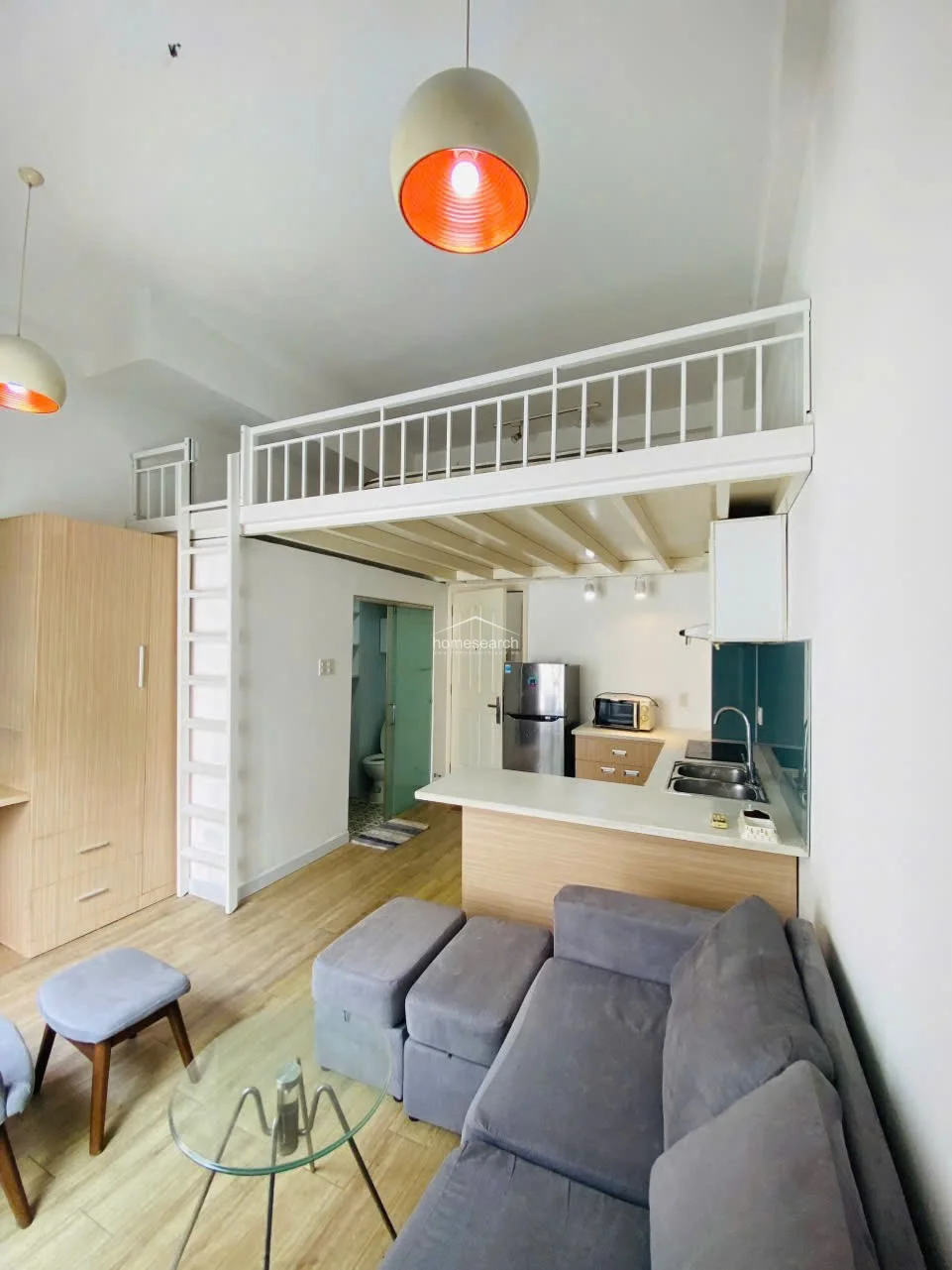 This shot captures the efficient layout of the studio, emphasizing the compact kitchen equipped with a stainless steel refrigerator and a modern sink. The living area is comfortably furnished with a grey sofa and ottoman, while the loft structure adds architectural interest and vertical storage potential.