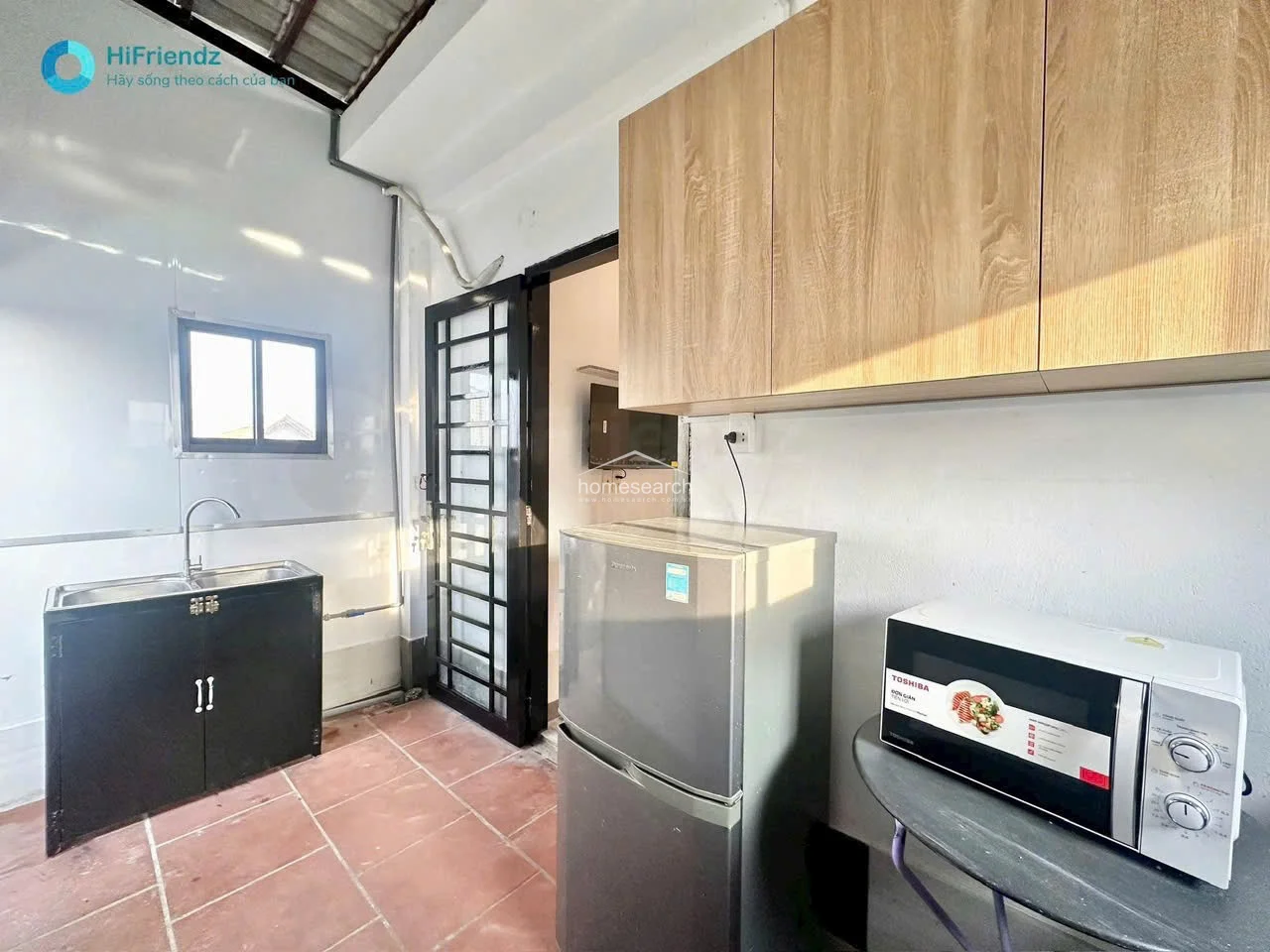 This kitchen area features a functional layout with a stainless steel sink, a compact refrigerator, and a microwave placed on a small stand. The space is finished with wood-grain upper cabinetry for storage and is connected to the bedroom through a doorway, with a window providing natural light.