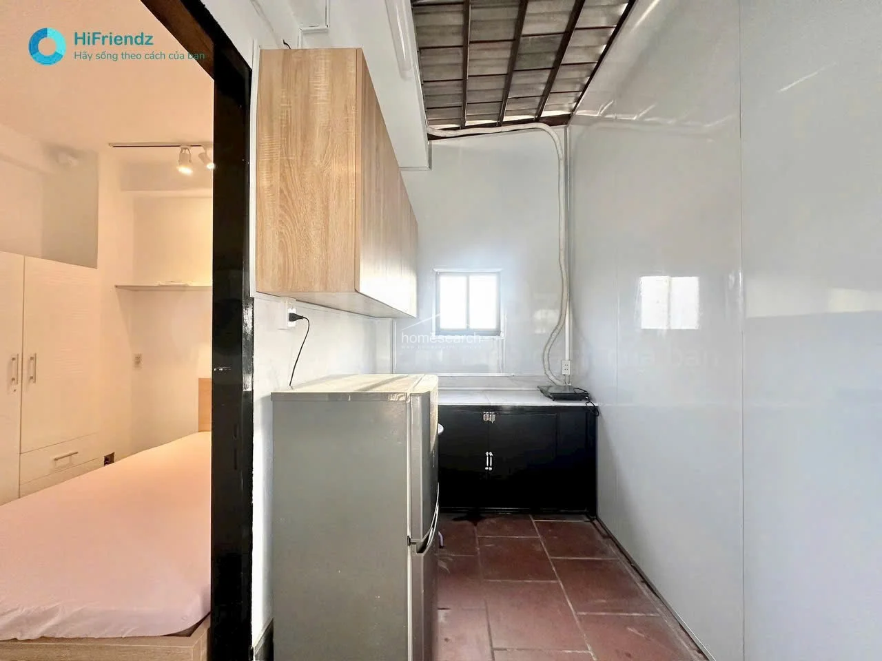 This view highlights the kitchen's connection to the bedroom, showcasing a clean, minimalist aesthetic with light-toned wood cabinets and white wall paneling. The area is illuminated by natural light from a window and features a simple, utilitarian design suitable for a small apartment.
