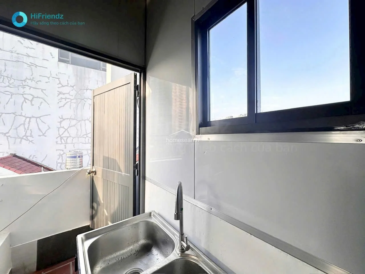This kitchen corner features a stainless steel sink set against a window that offers natural light and a view of the exterior. A door provides direct access to an outdoor balcony area, and the walls are finished with easy-to-clean white panels, indicating a practical and well-maintained space.