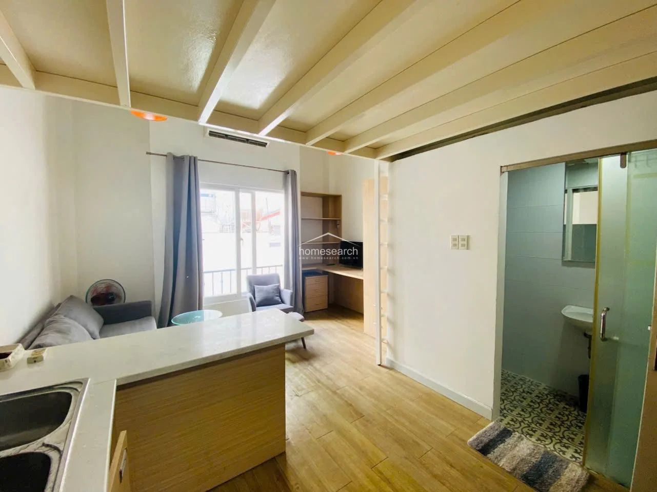 This studio apartment features a functional loft layout with light wood-look flooring and a clean, neutral color scheme. The space includes a built-in kitchen counter, a dedicated study nook with shelving, and a glass-enclosed bathroom with patterned tile flooring.