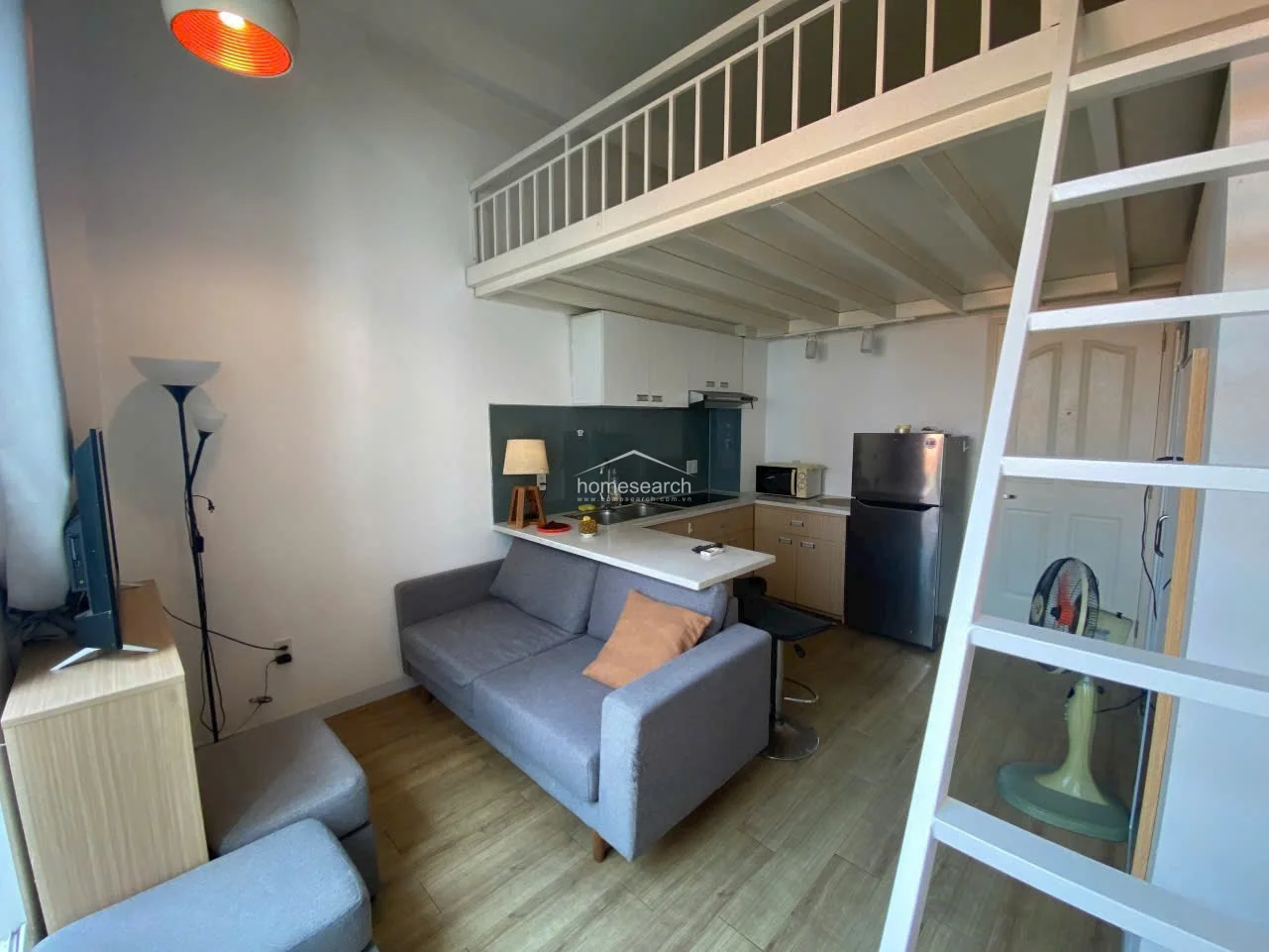 This view highlights the efficient open-plan living area, which includes a comfortable grey sofa and a compact kitchen equipped with a stainless steel refrigerator and microwave. The loft structure is accessed via a white ladder, maximizing the vertical space in this modern, well-maintained unit.