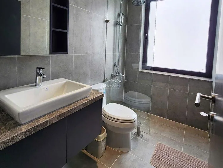 The bathroom maintains a contemporary aesthetic with large-format grey wall and floor tiles, paired with a floating vanity that features a dark cabinet and a stone-look countertop. It includes a modern white basin, a glass-enclosed shower area with a rainfall showerhead, and a frosted window that allows for natural light while maintaining privacy. The space is in excellent, well-maintained condition, reflecting a high-end, functional design.