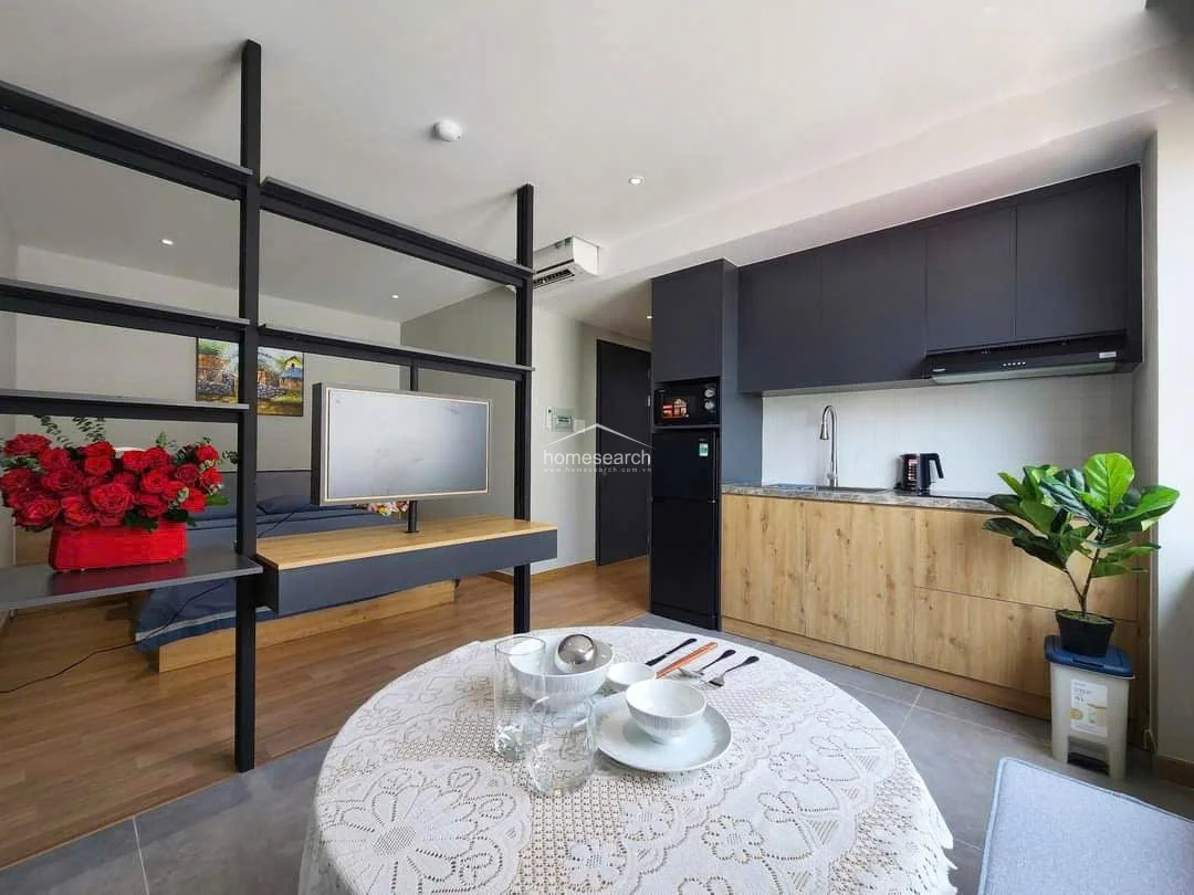 The kitchen area is efficiently designed with a linear layout, featuring dark cabinetry paired with light wood-tone lower cabinets for a contemporary contrast. It is equipped with essential appliances, including a built-in microwave and a compact refrigerator, and maintains a clean, minimalist look. The space flows directly into the dining area, which is anchored by a round table and a built-in bench seat, maximizing the functional use of the floor plan.