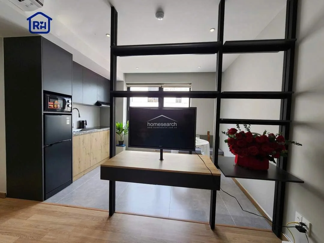 This view highlights the clever use of a floor-to-ceiling metal shelving partition that acts as both a room divider and a media console. The dark metal frame provides a striking architectural detail against the soft, neutral wall palette, while the integrated TV mount creates a seamless transition between the kitchen and the bedroom. The space feels bright and airy, benefiting from the natural light streaming in from the large window at the far end.