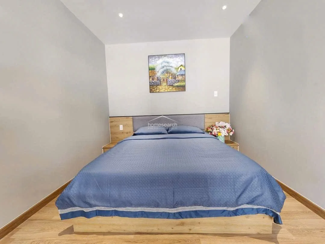 The bedroom area is cozy and well-defined, featuring a platform bed with a matching wood-tone headboard and integrated side tables. The soft blue bedding adds a splash of color to the neutral grey walls, while the wood flooring provides warmth to the sleeping quarters. The room is minimalist and uncluttered, emphasizing a high level of maintenance and a peaceful, restful atmosphere.