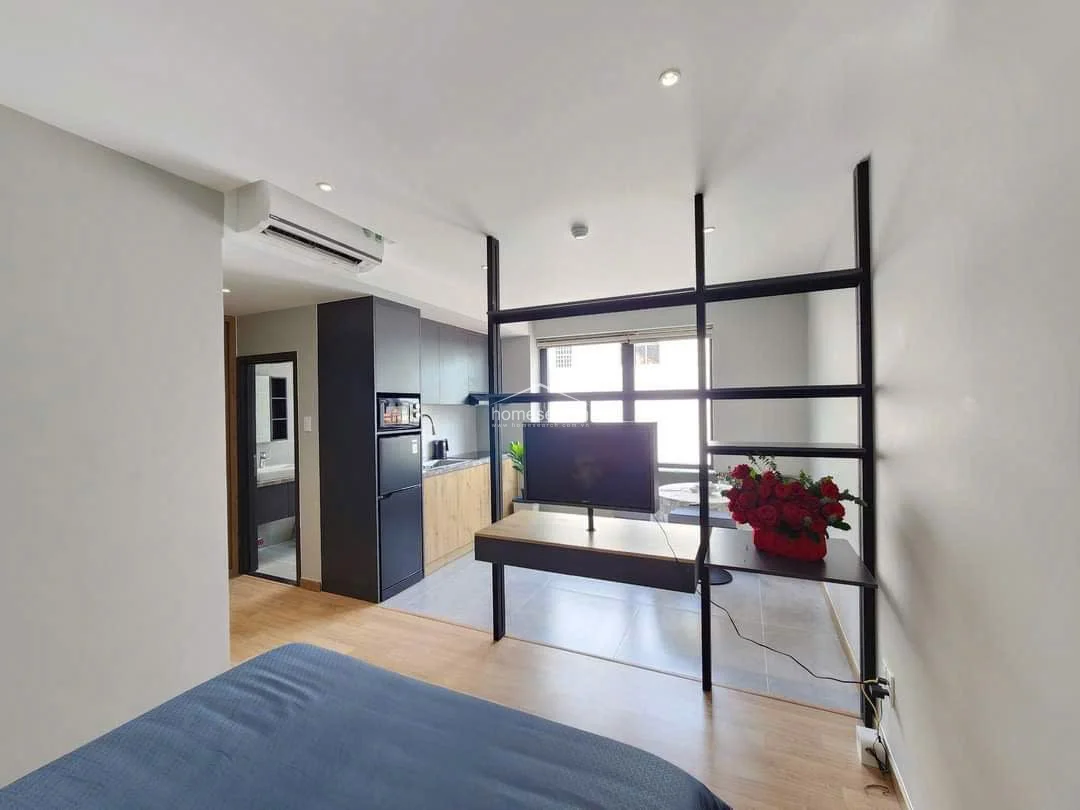 This perspective showcases the studio's overall flow, connecting the sleeping area, the central media partition, and the kitchen in a cohesive, open-plan design. A split-system AC unit is mounted above the kitchen entrance, ensuring climate control for the entire space, while a doorway leads into a private bathroom. The combination of recessed ceiling lighting and natural light creates a bright, inviting environment that feels both functional and stylish.