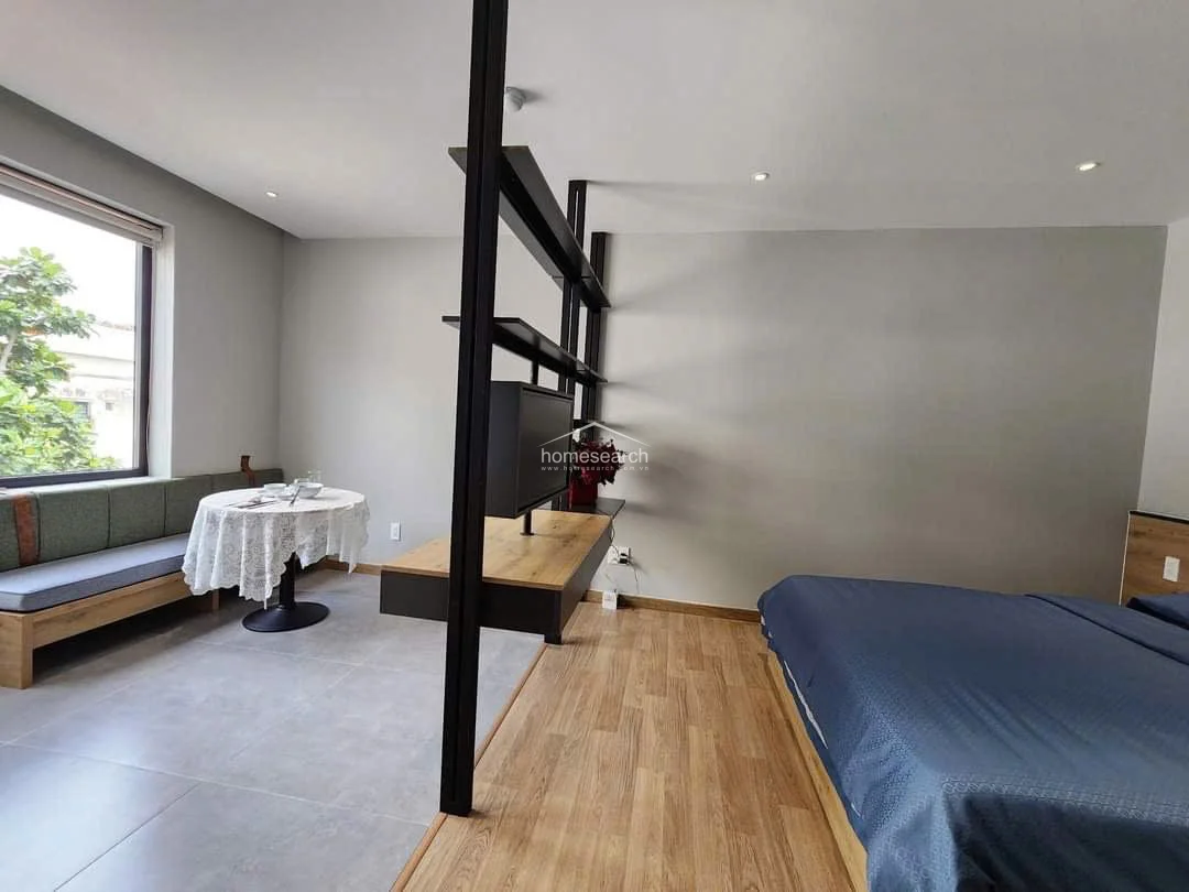 This studio apartment features a smart, open-concept layout that separates the sleeping area from the living space using a sleek, black metal shelving unit. The room is finished with a combination of light wood-look flooring in the bedroom and neutral grey tiles in the dining area, creating a modern and clean aesthetic. A large window provides ample natural light and a view of greenery, while the overall condition appears to be excellent and well-maintained.