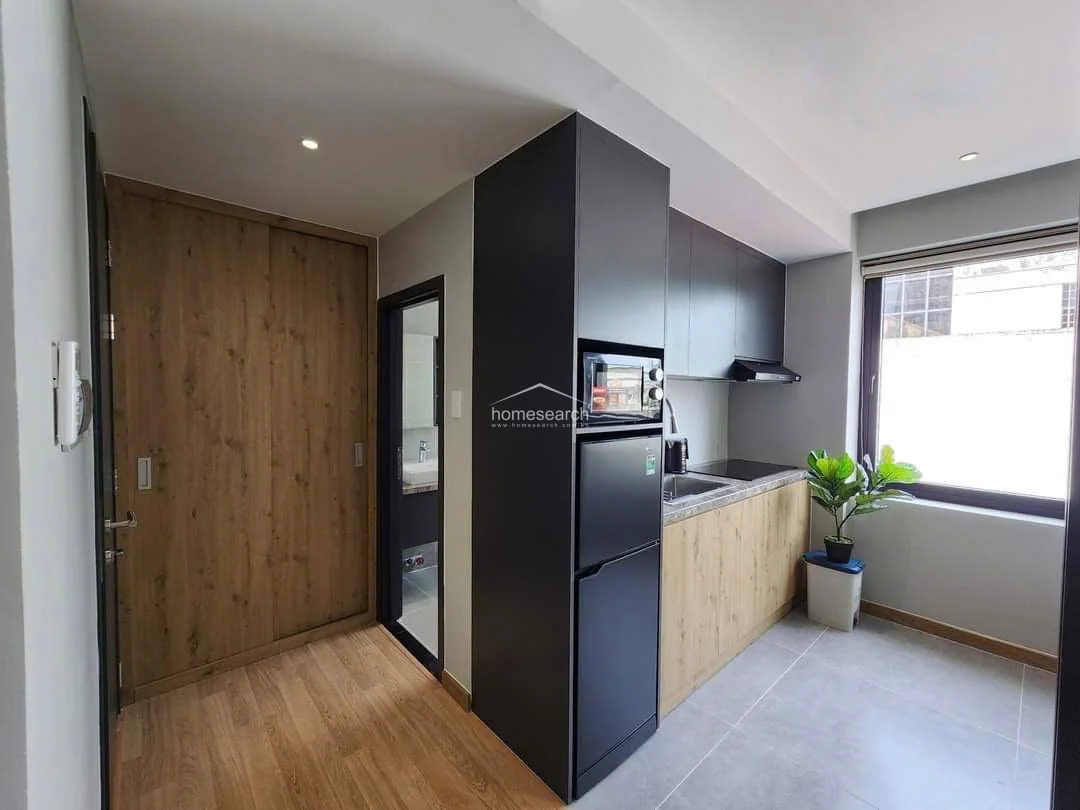 This compact, modern kitchenette features a sleek, two-tone design with matte black upper cabinetry and light wood-grain lower cabinets, complemented by grey tiled flooring and neutral walls. The space is efficiently laid out with an integrated microwave and refrigerator, while a large window provides natural light and a view of the adjacent building. The area flows directly into the entryway, which includes a matching wood-paneled closet and a doorway leading into the bathroom.