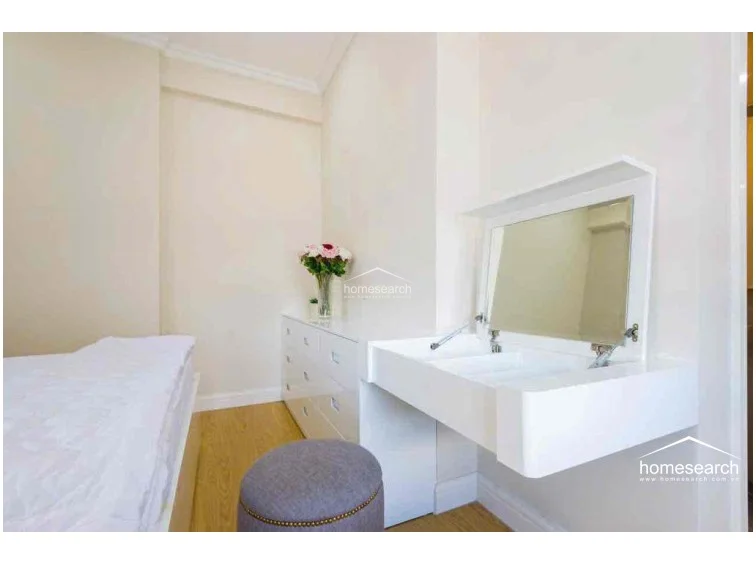 This compact bedroom features a clean, minimalist aesthetic with warm wood-tone flooring and crisp, off-white walls that enhance the sense of space. The room is efficiently furnished with a built-in white vanity featuring a flip-top mirror and a matching chest of drawers, providing practical storage and functionality. The overall condition appears excellent, with crown molding adding a subtle architectural detail to the well-maintained interior.