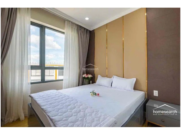 This bedroom benefits from ample natural light streaming through a large window, which is fitted with both sheer and heavy drapes for privacy and light control. The room is well-appointed with a comfortable bed, a matching bedside table, and a sophisticated wall treatment, reflecting a high level of maintenance and modern style.