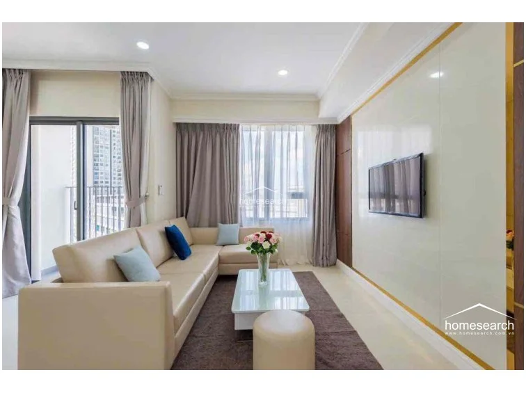 The living area is centered around a sleek wall-mounted television panel with gold accents, which adds a touch of sophistication to the room. The layout flows seamlessly toward a balcony through sliding glass doors, while the neutral decor and recessed lighting maintain a cohesive, modern aesthetic.