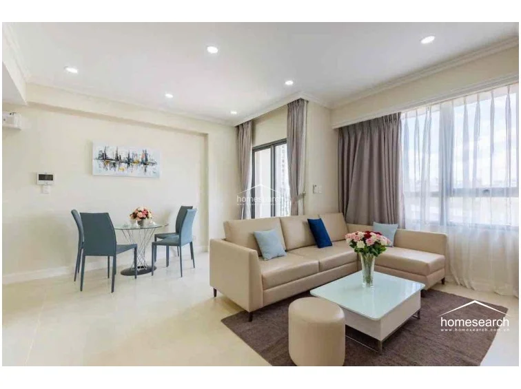 This open-concept living and dining area features light-colored tile flooring and a neutral color palette, creating a bright and airy atmosphere. The space is well-lit by recessed ceiling lights and large windows dressed in floor-to-ceiling curtains, with a glass-topped dining set and a modern L-shaped sofa providing a functional layout.