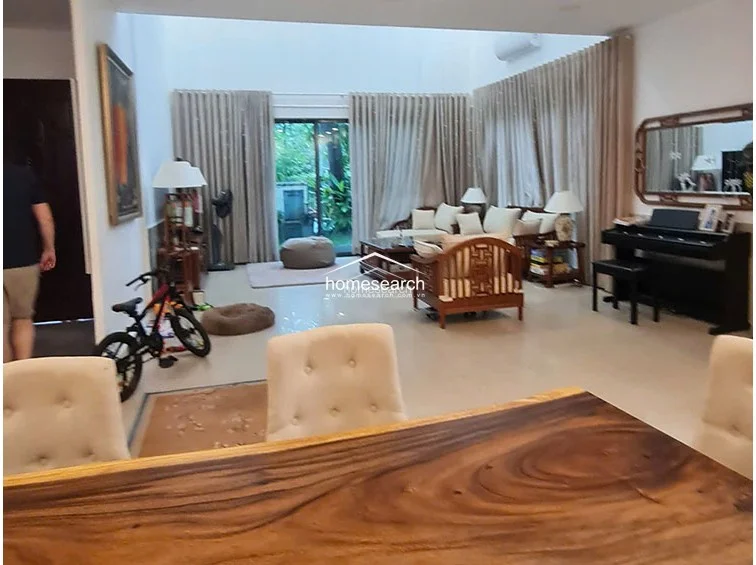The living area is designed with a comfortable seating arrangement centered around a wooden coffee table, positioned to overlook the outdoor garden through expansive floor-to-ceiling windows. The neutral color palette is accented by warm wood furniture and soft, floor-length curtains that provide privacy and texture. A piano is tucked into the corner, and the overall condition appears well-maintained with a focus on a relaxed, family-friendly layout.