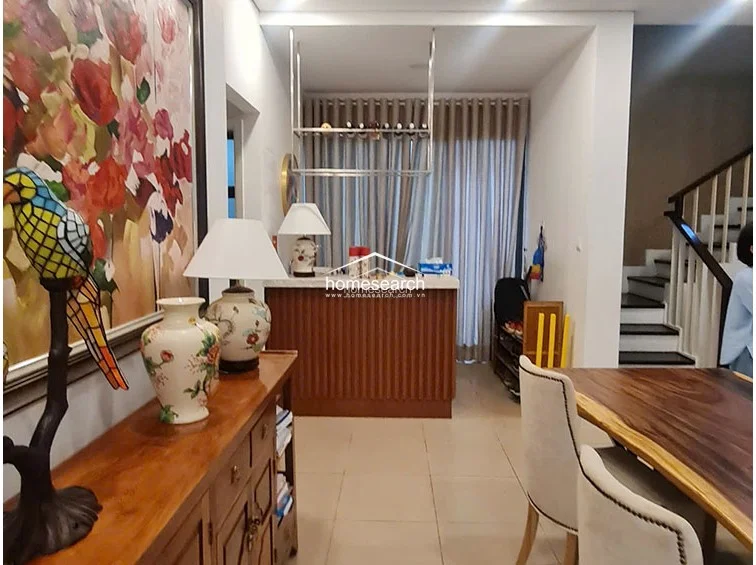 This view shows the dining area and a dedicated bar or serving counter, which adds a functional touch to the open floor plan. The space is decorated with a large, vibrant floral painting and decorative ceramic vases, adding personality to the neutral wall finishes. The wooden staircase with white balusters is visible to the right, maintaining the cohesive design language found throughout the residence.