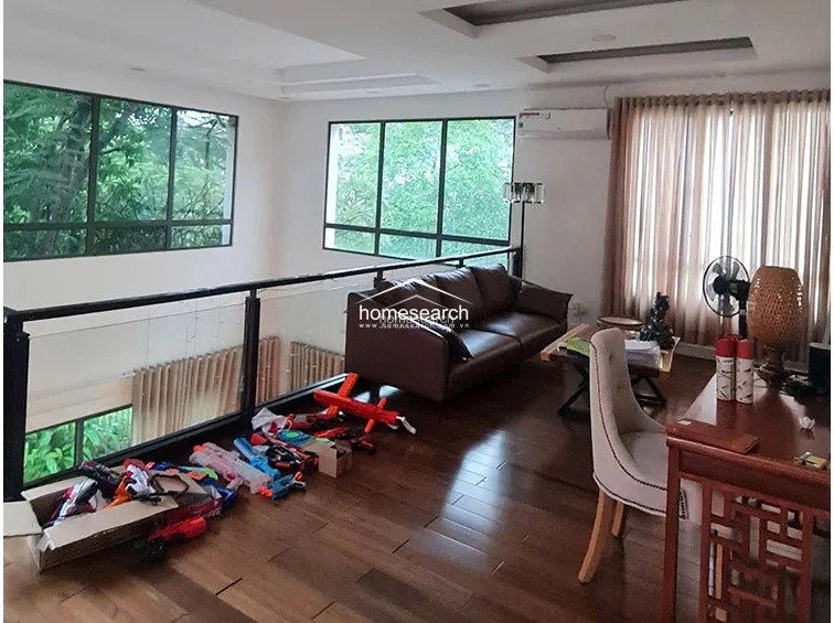 This mezzanine-style living area overlooks a lower level through a modern glass and metal railing, creating an airy, connected feel within the home. The space is furnished with a comfortable leather sofa and a wooden desk, accented by large windows that offer lush green views and ample natural light.