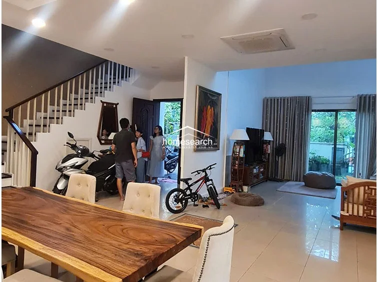 This open-concept living and dining area features a double-height ceiling that enhances the sense of space and natural light, complemented by large glass sliding doors leading to an outdoor area. The flooring is consistent with light-colored polished tiles, while the neutral walls and floor-to-ceiling curtains create a bright, airy atmosphere. A prominent wooden dining table serves as a centerpiece, and the space includes a ceiling-mounted AC unit and a staircase with a wooden handrail.