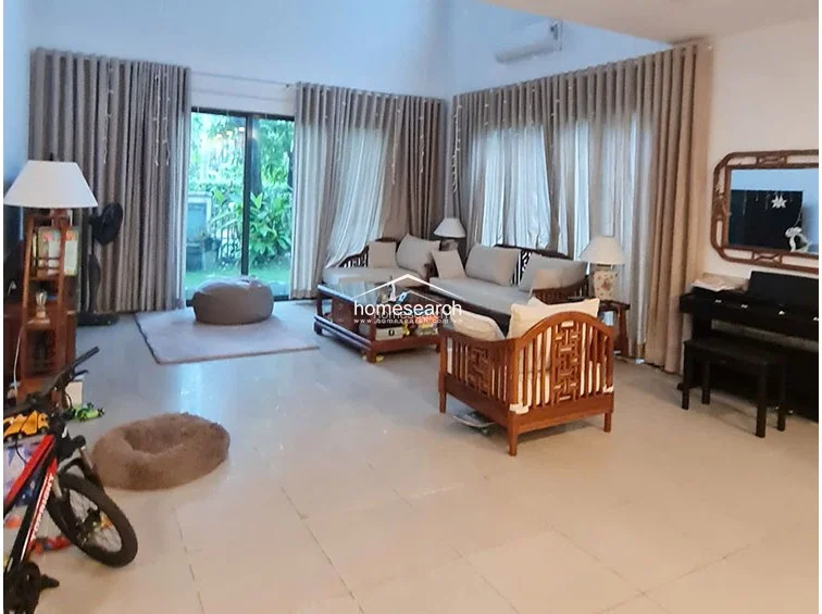 This view highlights the spacious, open-plan flow of the living room, emphasizing the high ceiling and the seamless transition to the outdoor space. The room is furnished with a mix of traditional wooden pieces and modern neutral upholstery, creating a balanced aesthetic. The polished tile flooring reflects the natural light, contributing to the clean and well-kept appearance of the home.