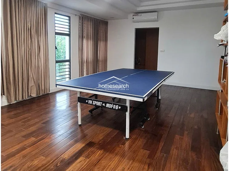 This spacious room features rich, dark-toned hardwood flooring and a minimalist white wall finish, currently utilized as a recreation area with a large table tennis set. The space is equipped with a wall-mounted AC unit and features floor-to-ceiling curtains that provide privacy while allowing for natural light through the side windows.
