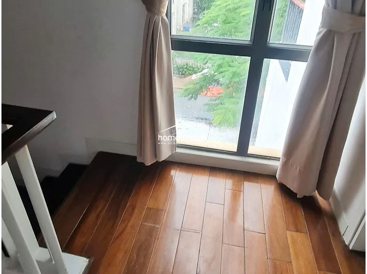This landing or transitional space features rich, polished wooden flooring and a large window draped with neutral curtains that allow for natural light. The area connects to a staircase with a simple white railing, creating an open and airy feel with a view of the outdoor foliage.