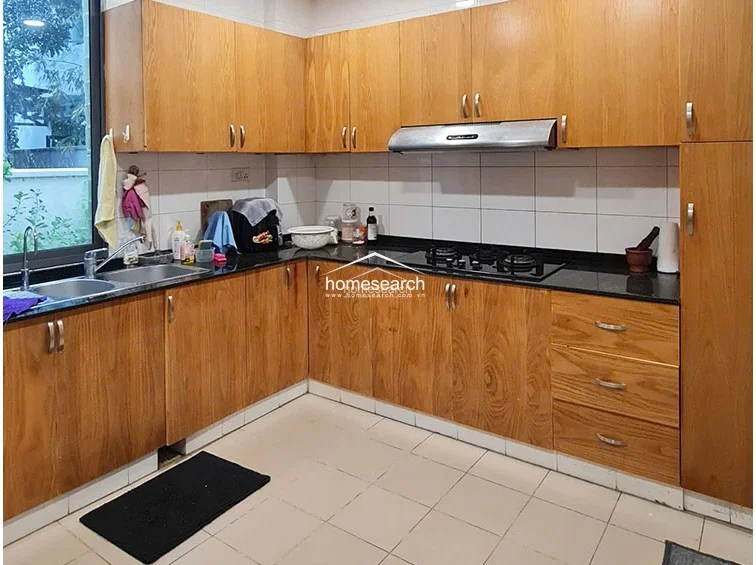 This kitchen layout maintains a consistent aesthetic with wooden cabinets and black countertops, featuring a stainless steel range hood and a gas cooktop. The space is functional and well-lit by the adjacent window, with white tiled walls that offer a clean, easy-to-maintain finish.