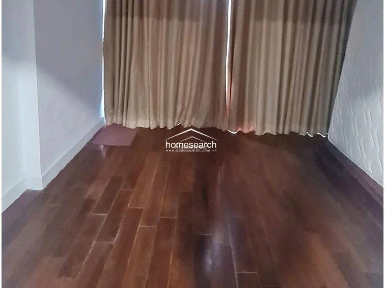 This room is characterized by warm, polished wood flooring and floor-to-ceiling curtains that provide privacy and light control. The walls are painted a simple, neutral white, offering a blank canvas that is well-maintained and ready for furniture.